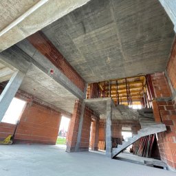 Perfekt-Haus Dawid Kokot - Surowy stan domu murowanego w trakcie budowy, widoczne ściany z czerwonej cegły, betonowy strop, schody i słupy, żółty wózek paletowy w tle, lekki nieporządek na budowie.