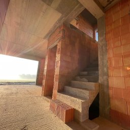 Perfekt-Haus Dawid Kokot - Surowy stan domu z cegły ceramicznej, widoczne betonowe schody i wschodzące słońce przez otwór okienny, z paletą cegieł na podłodze.