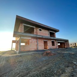 Perfekt-Haus Dawid Kokot - Dwukondygnacyjny dom w stanie surowym otwartym z czerwonej cegły, widoczny na tle błękitnego nieba i mglistego krajobrazu, z przyczepą kempingową w tle, na nierównym terenie.