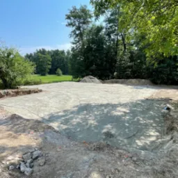 Świeżo wylana płyta fundamentowa na przygotowanym terenie, widoczne ślady opon i cień drzew, w tle zieleń drzew i błękitne niebo.