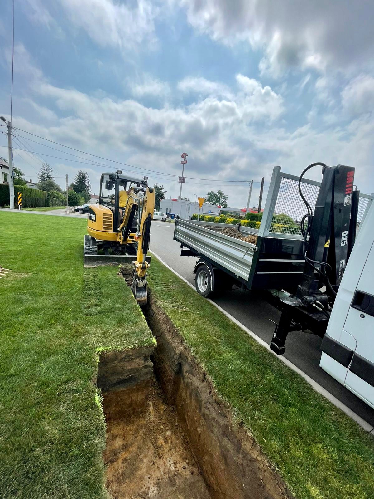Żółta mini koparka CAT wykonuje wąski wykop w trawiastym terenie wzdłuż drogi, obok zaparkowana przyczepa z wywrotką i żurawiem załadowczym.