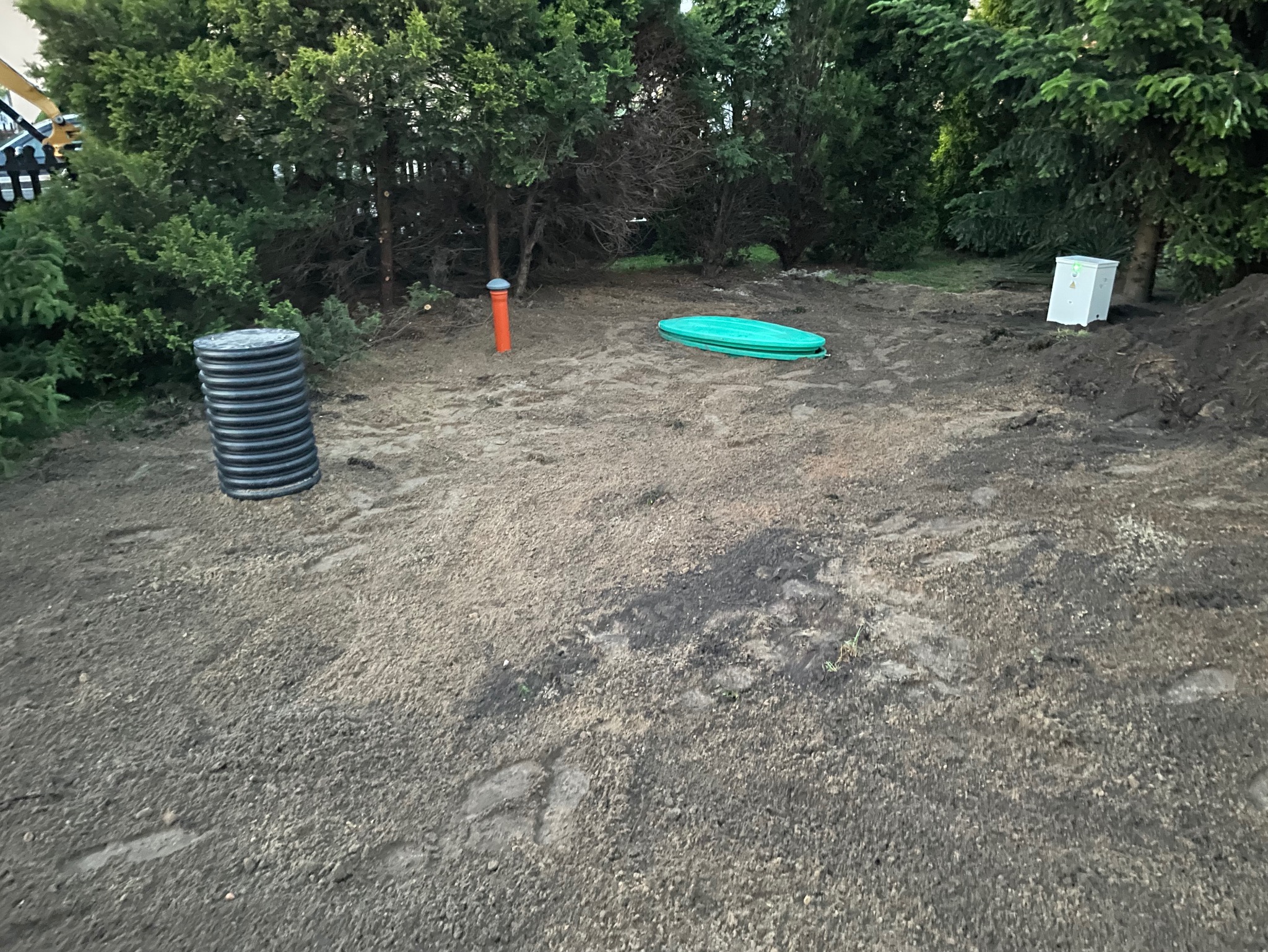 Teren po montażu przydomowej oczyszczalni ścieków: widoczne elementy instalacji, w tym czarna rura karbowana, pomarańczowy odpowietrznik, zielona pokrywa włazu oraz biała skrzynka elektryczna,...