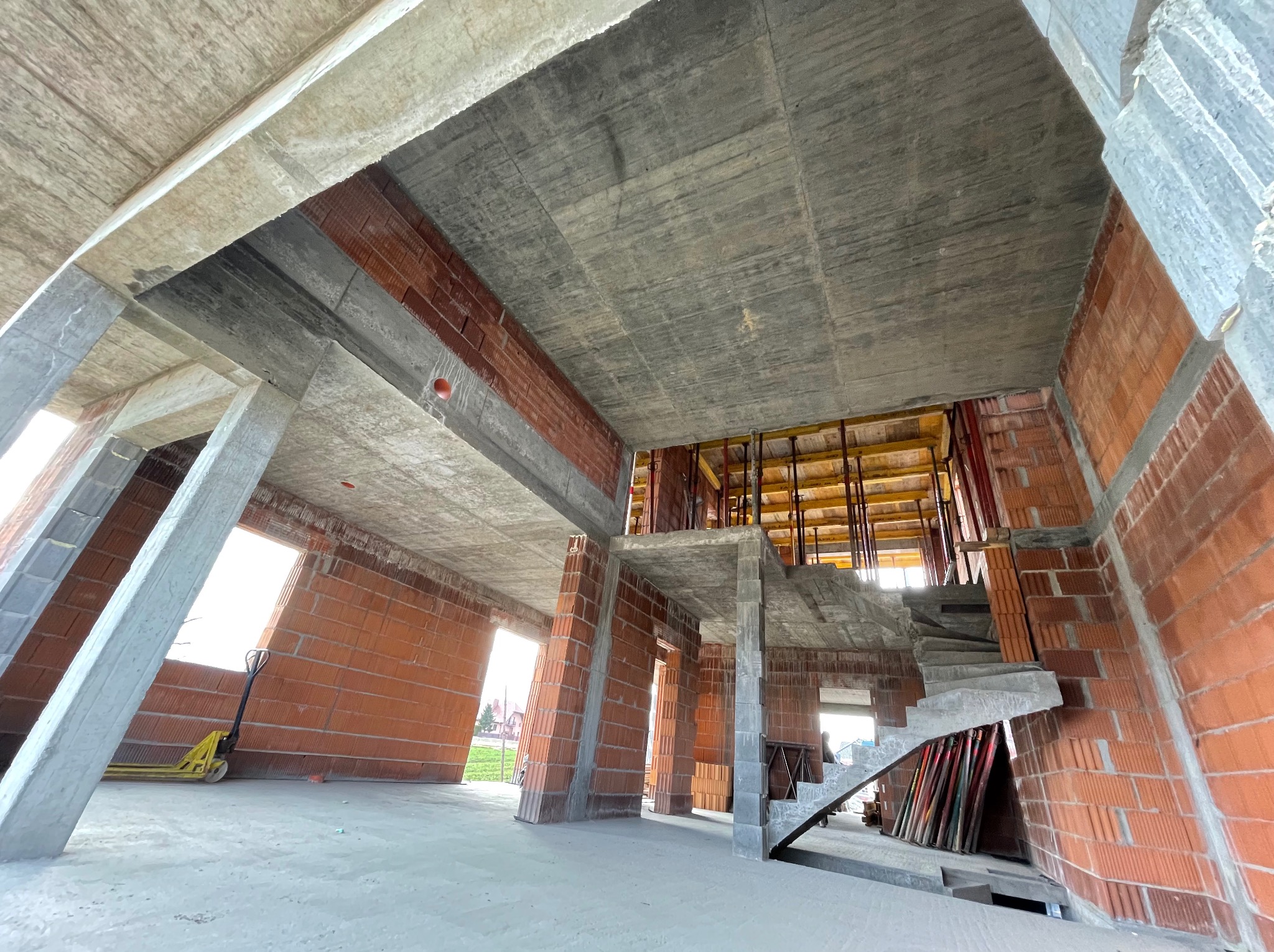 Surowy stan domu murowanego w trakcie budowy, widoczne ściany z czerwonej cegły, betonowy strop, schody i słupy, żółty wózek paletowy w tle, lekki nieporządek na budowie.