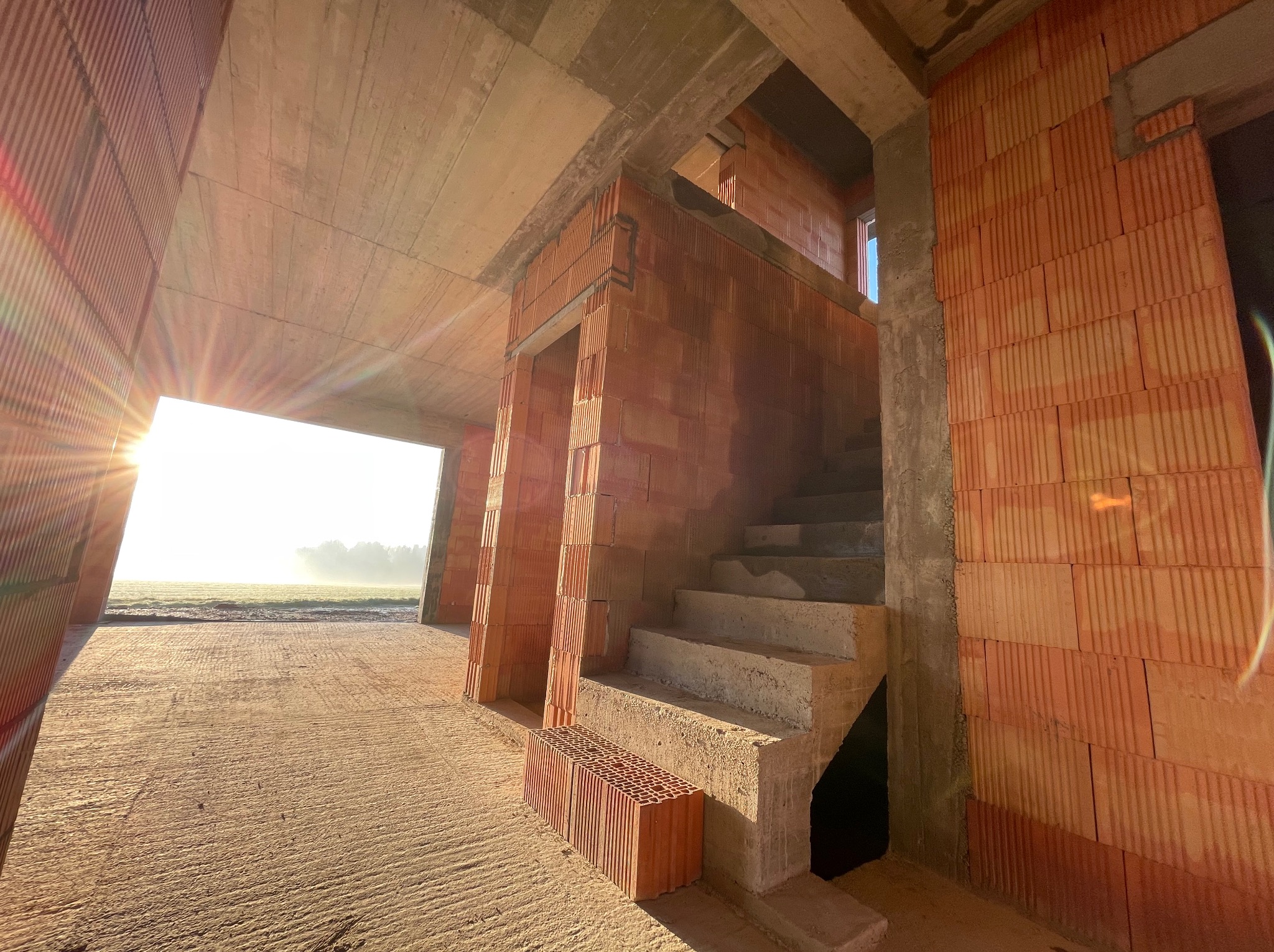 Surowy stan domu z cegły ceramicznej, widoczne betonowe schody i wschodzące słońce przez otwór okienny, z paletą cegieł na podłodze.