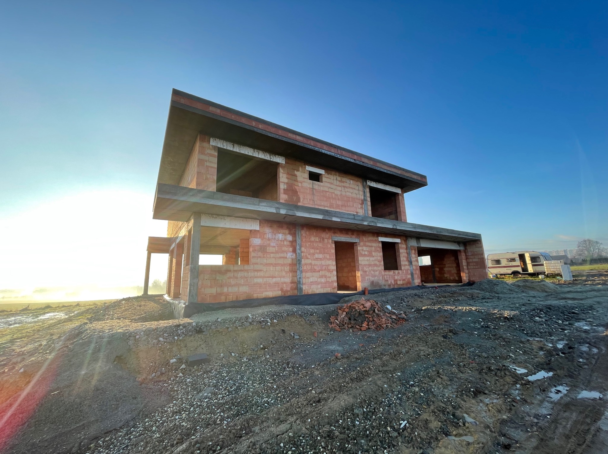 Dwukondygnacyjny dom w stanie surowym otwartym z czerwonej cegły, widoczny na tle błękitnego nieba i mglistego krajobrazu, z przyczepą kempingową w tle, na nierównym terenie.