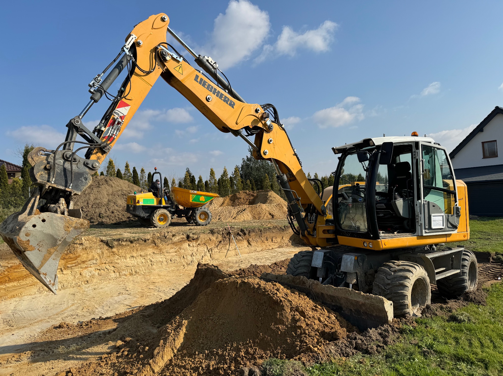 Żółta koparka Liebherr w trakcie prac ziemnych, widoczny wykop z warstwami gleby oraz wywrotka w tle, błękitne niebo.