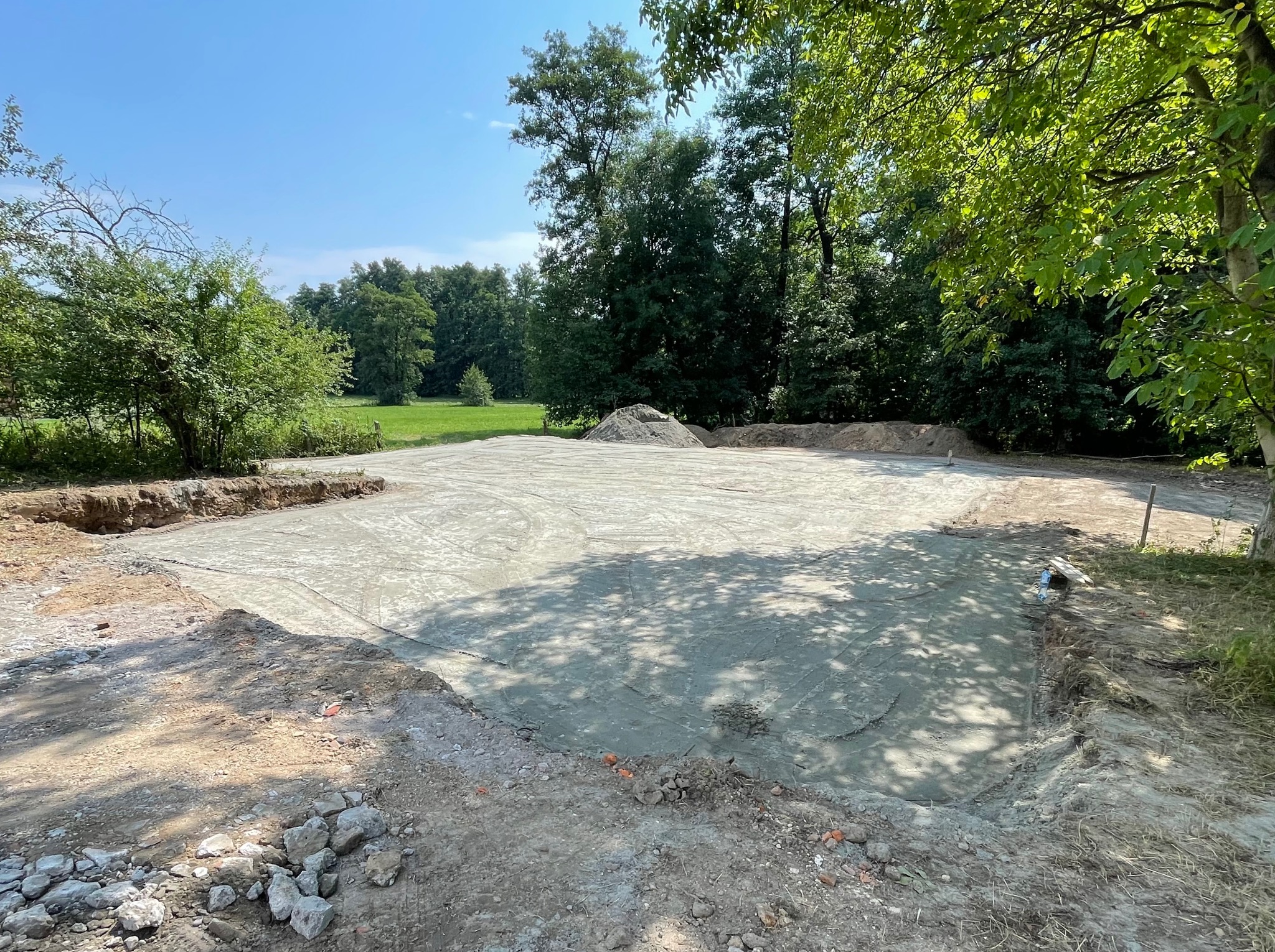 Świeżo wylana płyta fundamentowa na przygotowanym terenie, widoczne ślady opon i cień drzew, w tle zieleń drzew i błękitne niebo.