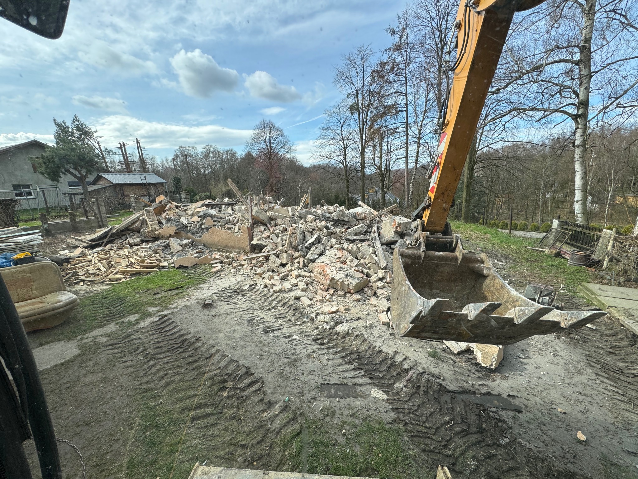 Żółta koparka z łyżką na pierwszym planie, w tle gruzowisko po wyburzonym budynku gospodarczym, widoczne ślady opon na błotnistym terenie.