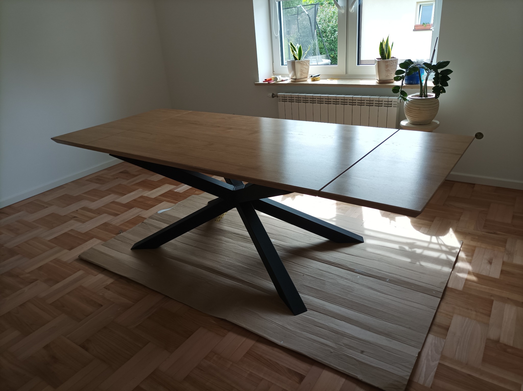Rozkładany stół z jasnego drewna na czarnych, geometrycznych nogach, zabezpieczony kartonem podczas montażu, na parkiecie w jodełkę, z oknem i roślinami w tle.
