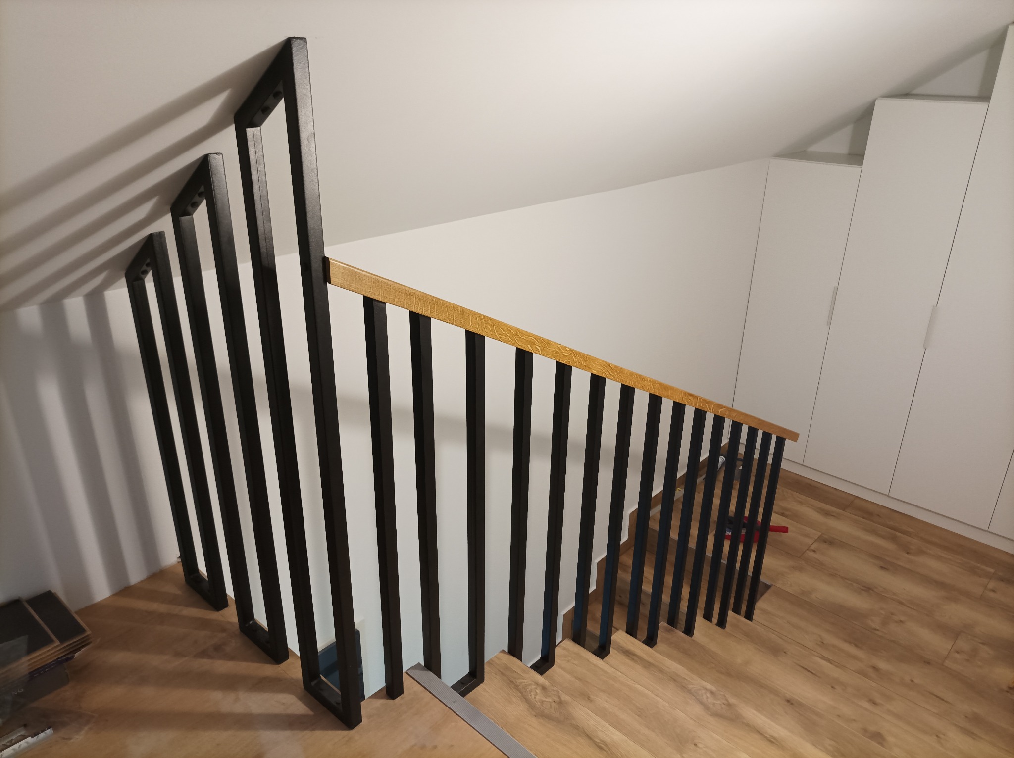 Nowoczesne schody drewniane z czarną, metalową balustradą o prostych, geometrycznych kształtach w jasnym wnętrzu z zabudową szaf w skosie.