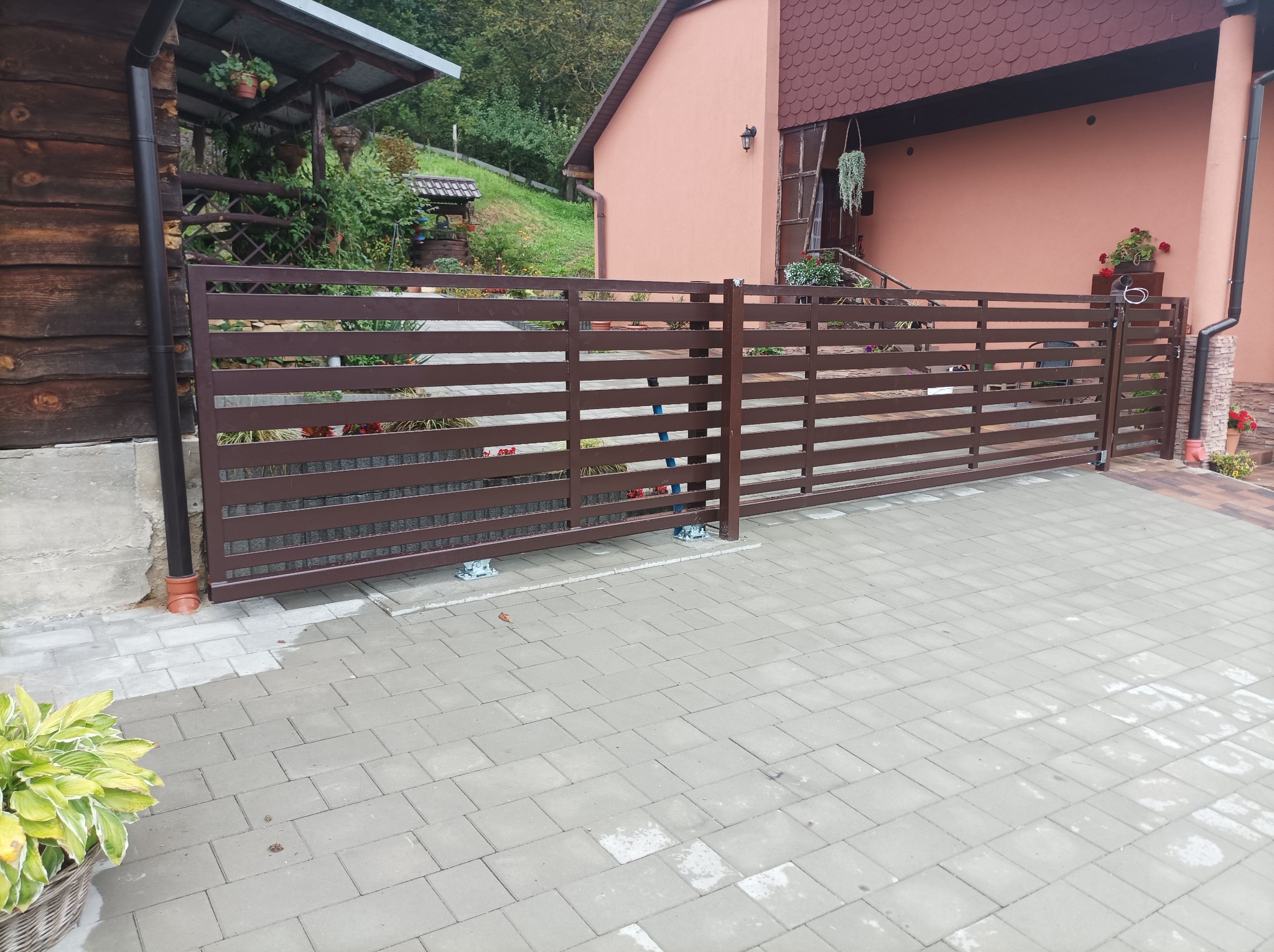 Brązowa, przesuwna brama z poziomych profili stalowych zamontowana przed domem z różową elewacją i kostką brukową na podjeździe, widoczne mechanizmy jezdne.