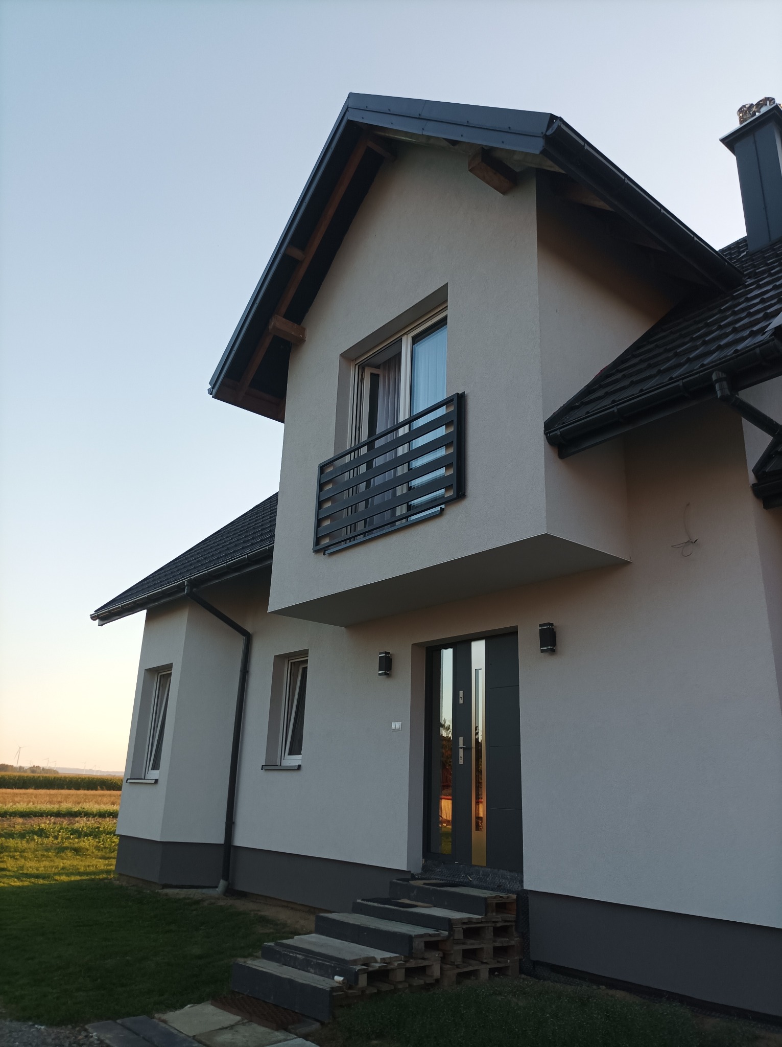 Nowoczesny dom jednorodzinny z balkonem i szarą stolarką okienną, widok z zewnątrz na tle pola i zachodzącego słońca.