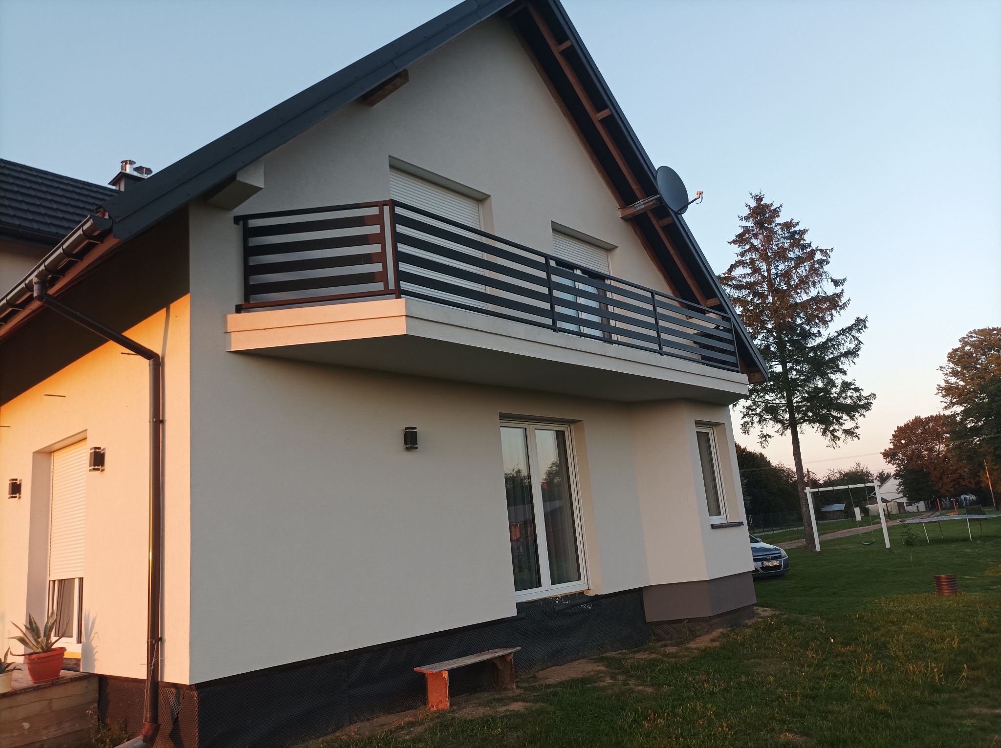 Nowoczesny dom jednorodzinny z balkonem o stalowej balustradzie z poziomymi poprzeczkami, oświetlony ciepłym światłem zachodzącego słońca, widok z boku na tle zielonego trawnika i drzew.