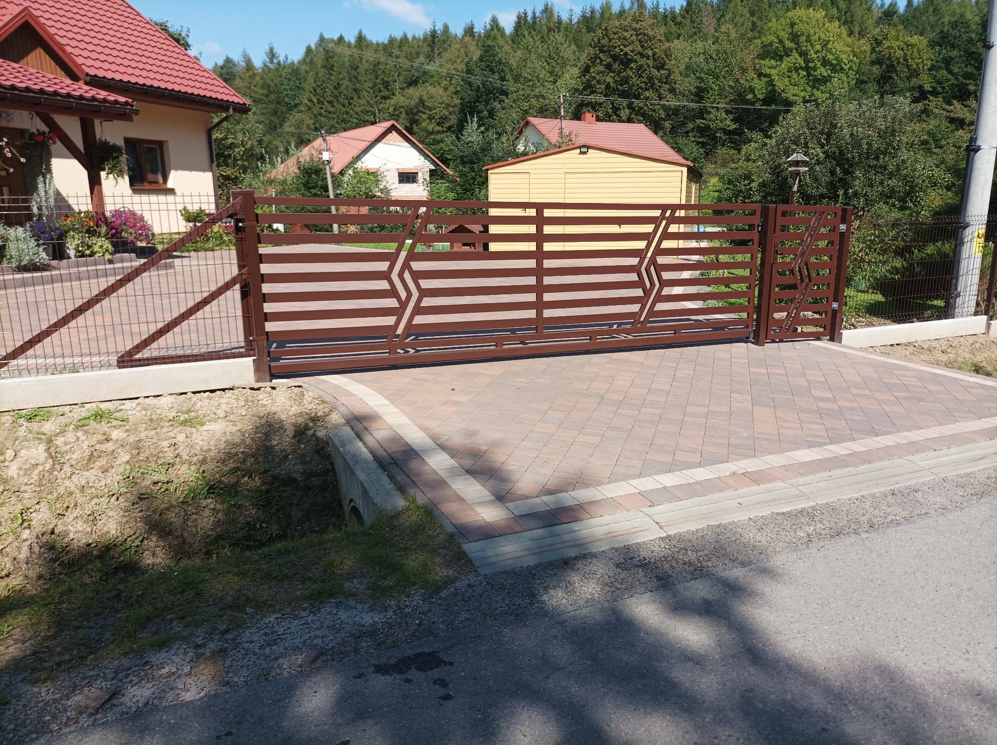 Brązowa, metalowa brama przesuwna z poziomymi szczeblami i geometrycznymi wzorami, zamontowana na posesji z kostką brukową, w tle domy i drzewa.