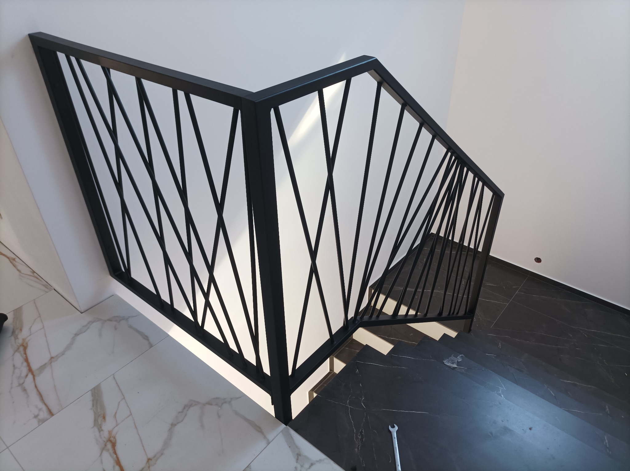 Nowoczesna, czarna balustrada schodowa z geometrycznym wzorem, widok z góry na marmurowe schody i podłogę, klucz płaski leżący na posadzce.