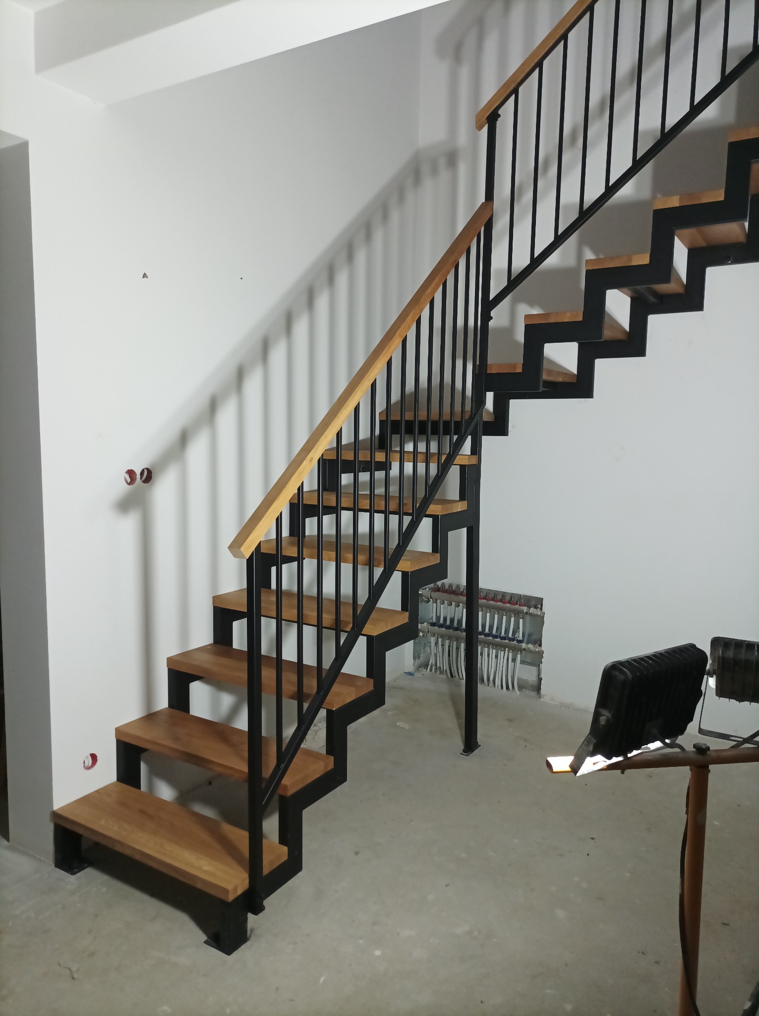 Nowoczesne schody wewnętrzne z drewnianymi stopniami i czarną metalową konstrukcją, widoczne instalacje grzewcze w ścianie, oświetlenie budowlane.