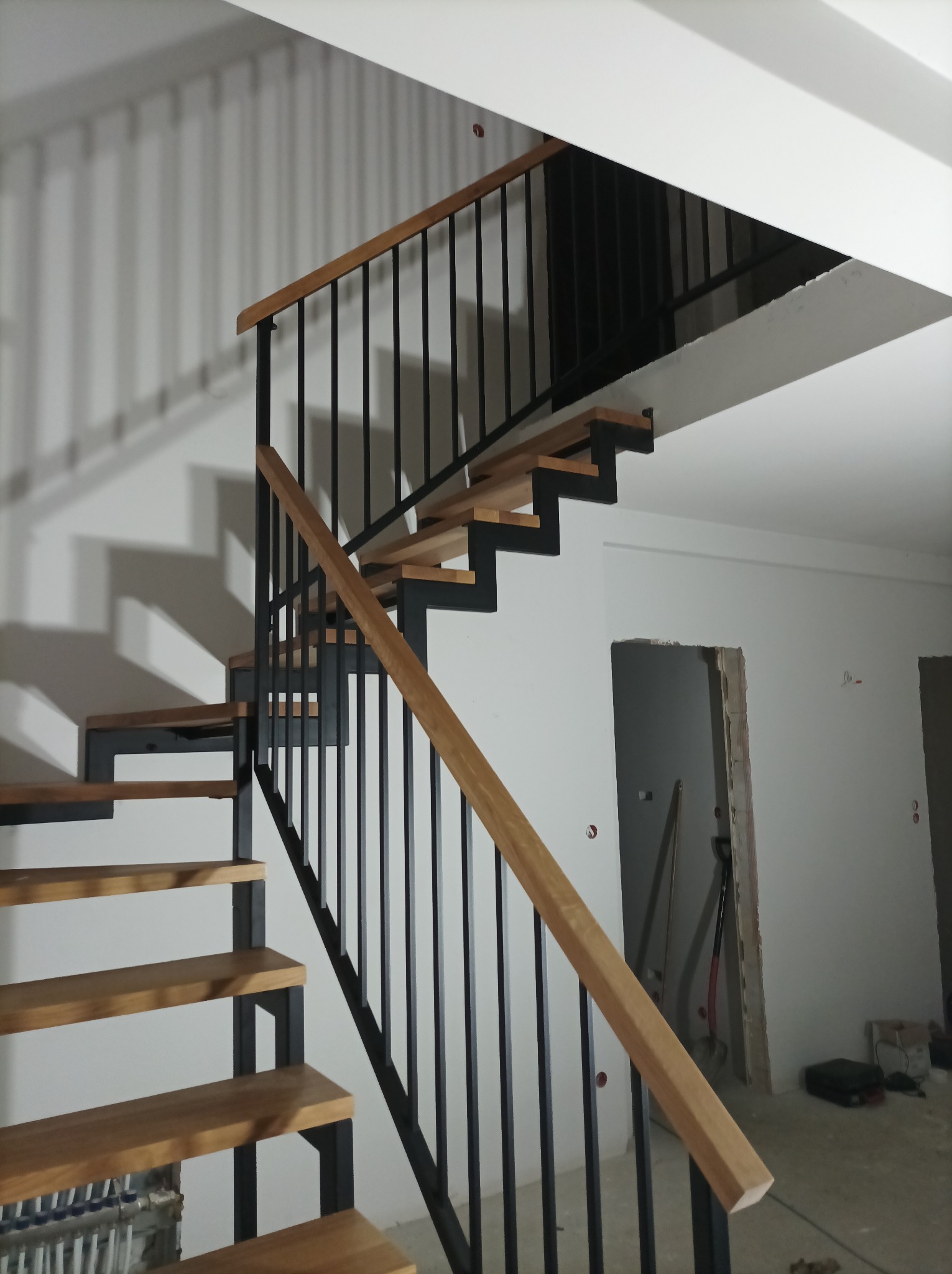 Nowoczesne schody z drewnianymi stopniami i czarną metalową konstrukcją, widoczne w surowym wnętrzu z niedokończonymi ścianami i otworami drzwiowymi.