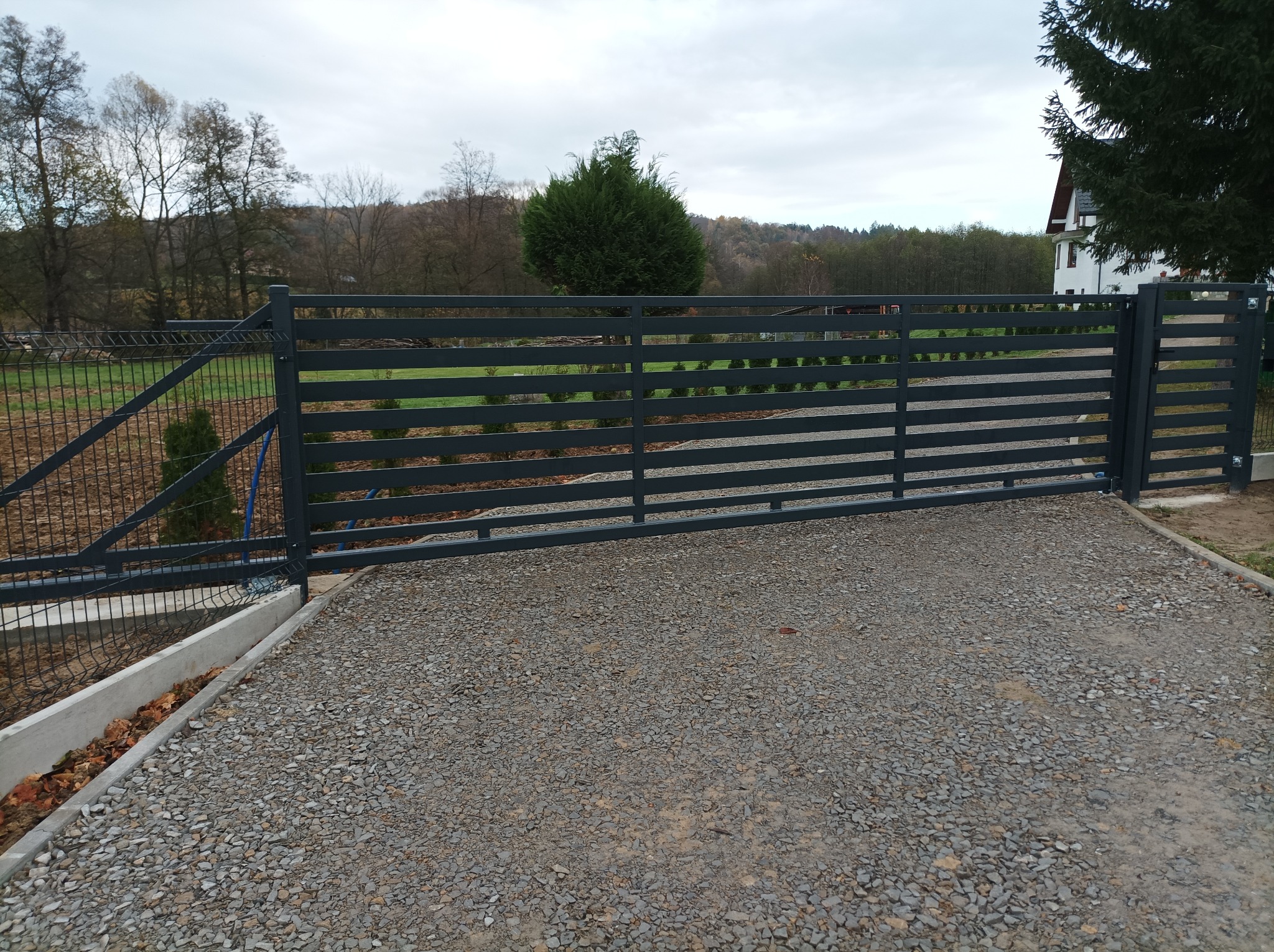 Nowoczesna, pozioma brama przesuwna w kolorze grafitowym na posesji z wysypanym żwirem podjazdem, widoczna siatka ogrodzeniowa i dom w tle.