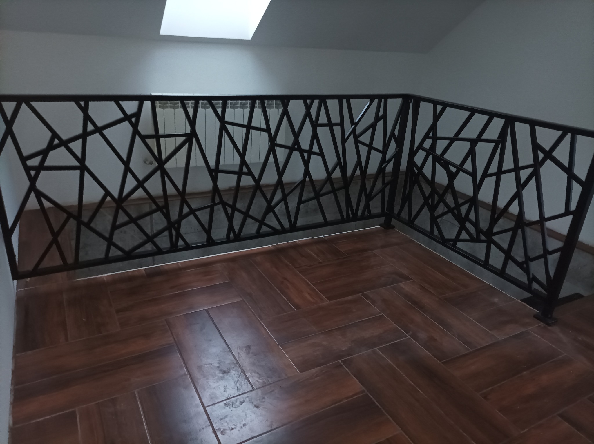 Ażurowa balustrada ze stali w kolorze czarnym, z motywem nieregularnych linii, na tle białej ściany i podłogi z brązowych płytek imitujących drewno, widoczny fragment grzejnika.