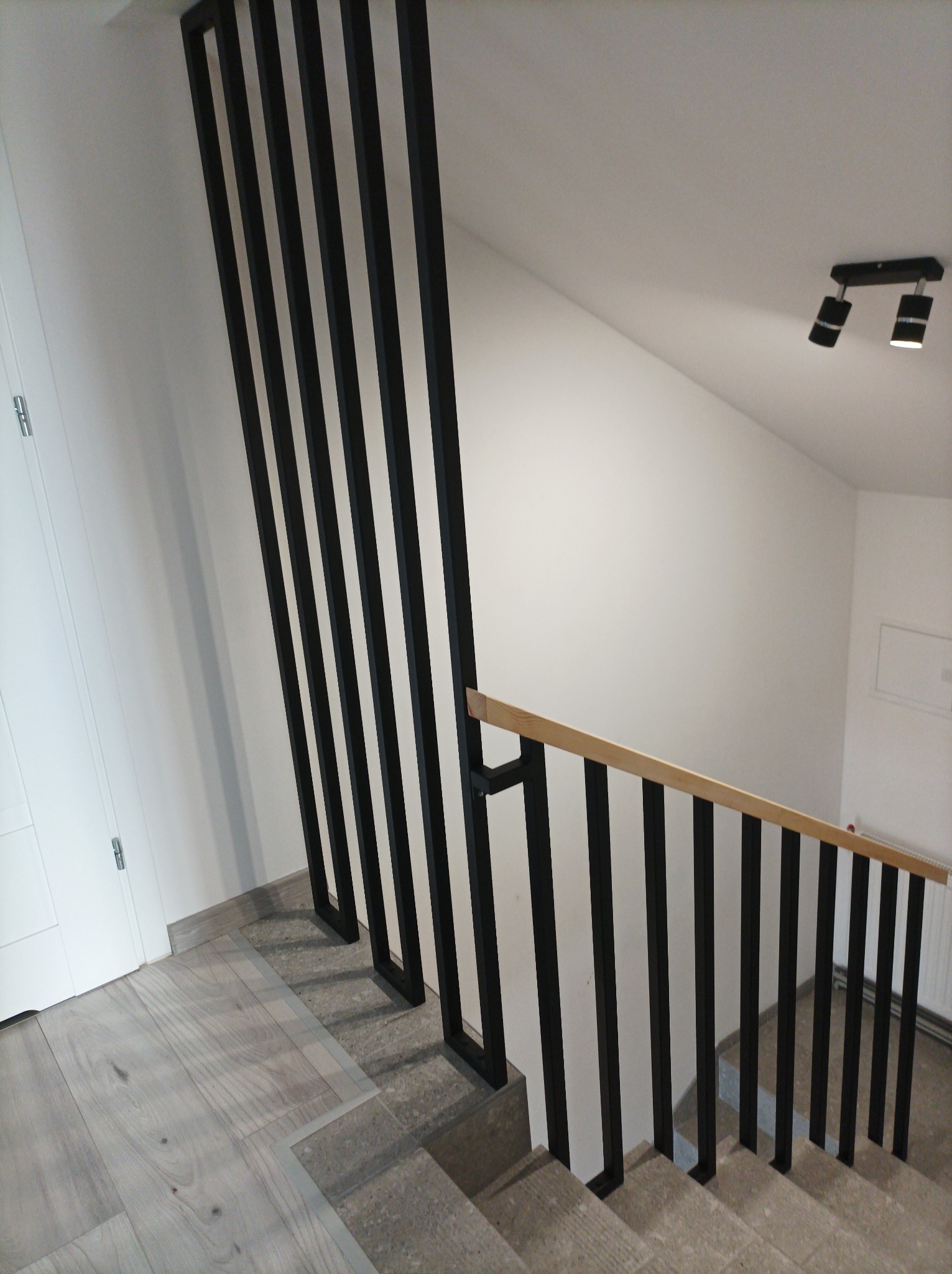 Nowoczesne schody z czarną, metalową balustradą o pionowych szczeblach i drewnianym poręczą, widok z góry na dół, jasne wnętrze z oświetleniem punktowym.