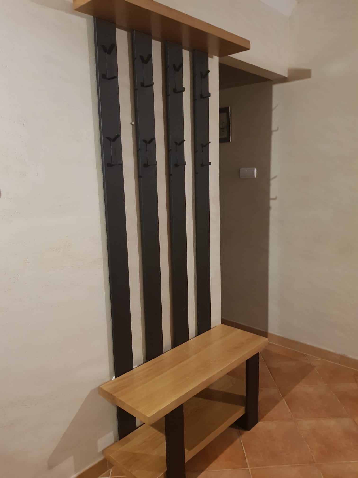 Nowoczesny wieszak z siedziskiem w przedpokoju, czarne metalowe elementy i drewno w odcieniach brązu, widoczne haki na ubrania.