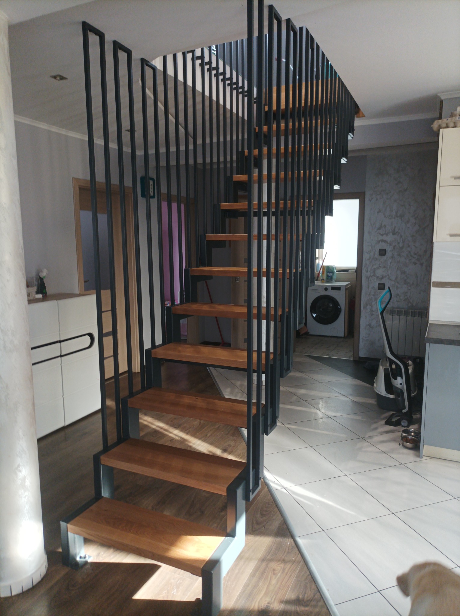 Nowoczesne schody z drewnianymi stopniami i czarną, stalową konstrukcją w jasnym wnętrzu domu, widoczna pralka w tle.