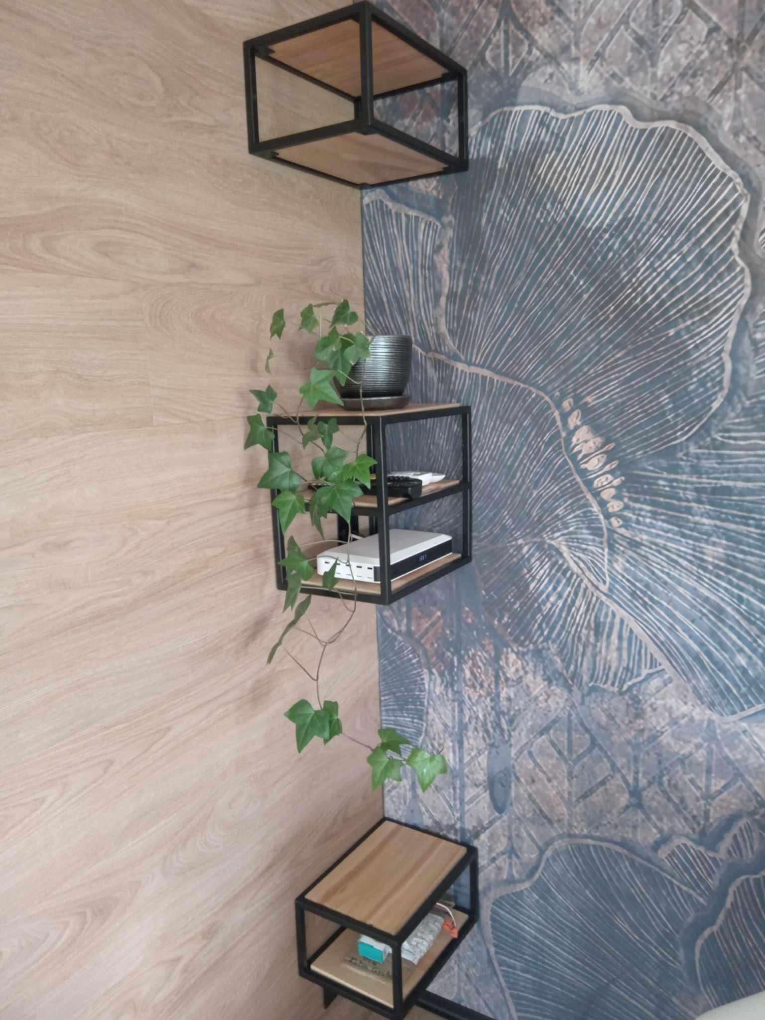 Trzy modułowe półki stalowe z drewnianymi blatami zamontowane w rogu pokoju z tapetą w motywy roślinne, na środkowej półce doniczka z bluszczem, dekoder i pilot.