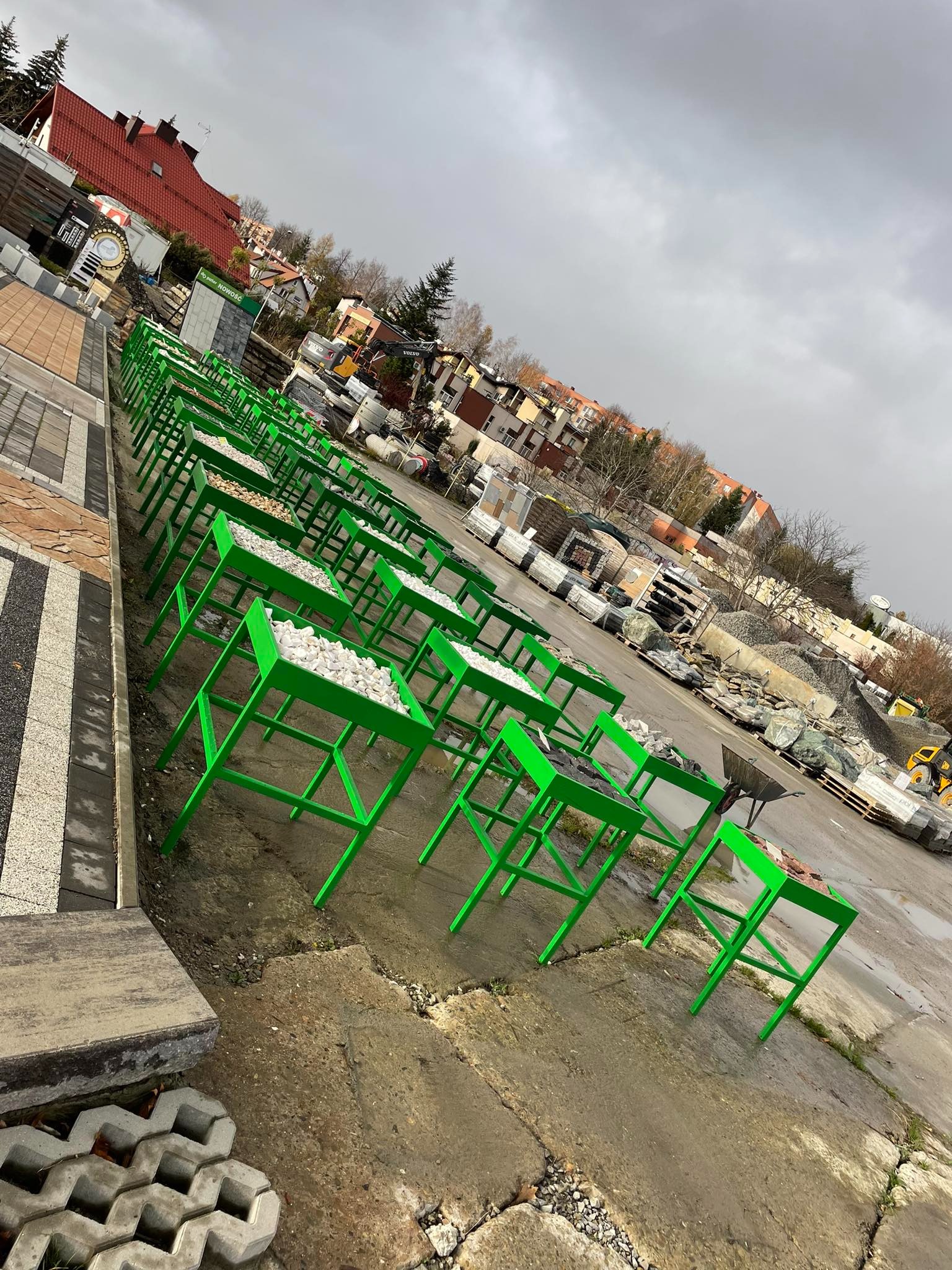 Ekspozycja próbek kamieni ozdobnych w zielonych, metalowych stelażach na placu budowlanym, w tle materiały budowlane i zabudowa miejska.