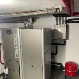 DDC Arkadiusz Obszyński - Szary piec DEFRO z podłączoną instalacją CO w kotłowni. Czerwony zbiornik wyrównawczy i orurowanie pod sufitem. Widok na nowoczesny system grzewczy.