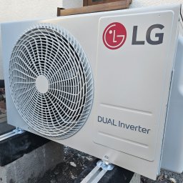 DDC Arkadiusz Obszyński - Zewnętrzna jednostka klimatyzacji LG Dual Inverter zamontowana na betonowych blokach przy ścianie budynku. Widoczne rury instalacyjne i wentylator jednostki.