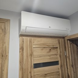 DDC Arkadiusz Obszyński - Biały klimatyzator LG Dual Inverter zamontowany nad drewnianą futryną drzwiową w jasnym wnętrzu. Widoczne detale urządzenia i fragmenty pomieszczenia.