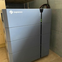 DDC Arkadiusz Obszyński - Szary akumulator energii Solplanet z diodami LED stanu na tle beżowych płytek i drewnianej półki. Widoczne przewody. Minimalistyczny design.