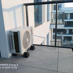 DDC Arkadiusz Obszyński - Biała jednostka zewnętrzna klimatyzacji Rotenso na balkonie nowoczesnego apartamentu, widok przez szklaną balustradę na sąsiedni budynek. Montaż na gumowych podkładkach antywibracyjnych.