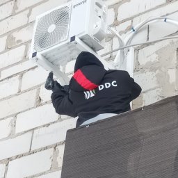 DDC Arkadiusz Obszyński - Montaż klimatyzatora Gorenje na białej ścianie z cegły. Instalator w czarnej kurtce z wkrętarką. Widoczne przewody i elementy montażowe.