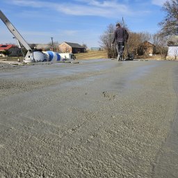 DDC Arkadiusz Obszyński - Wylewanie stropu z betonomieszarki na budowie domu jednorodzinnego. Pracownik wyrównuje powierzchnię. W tle zabudowa wiejska i błękitne niebo.