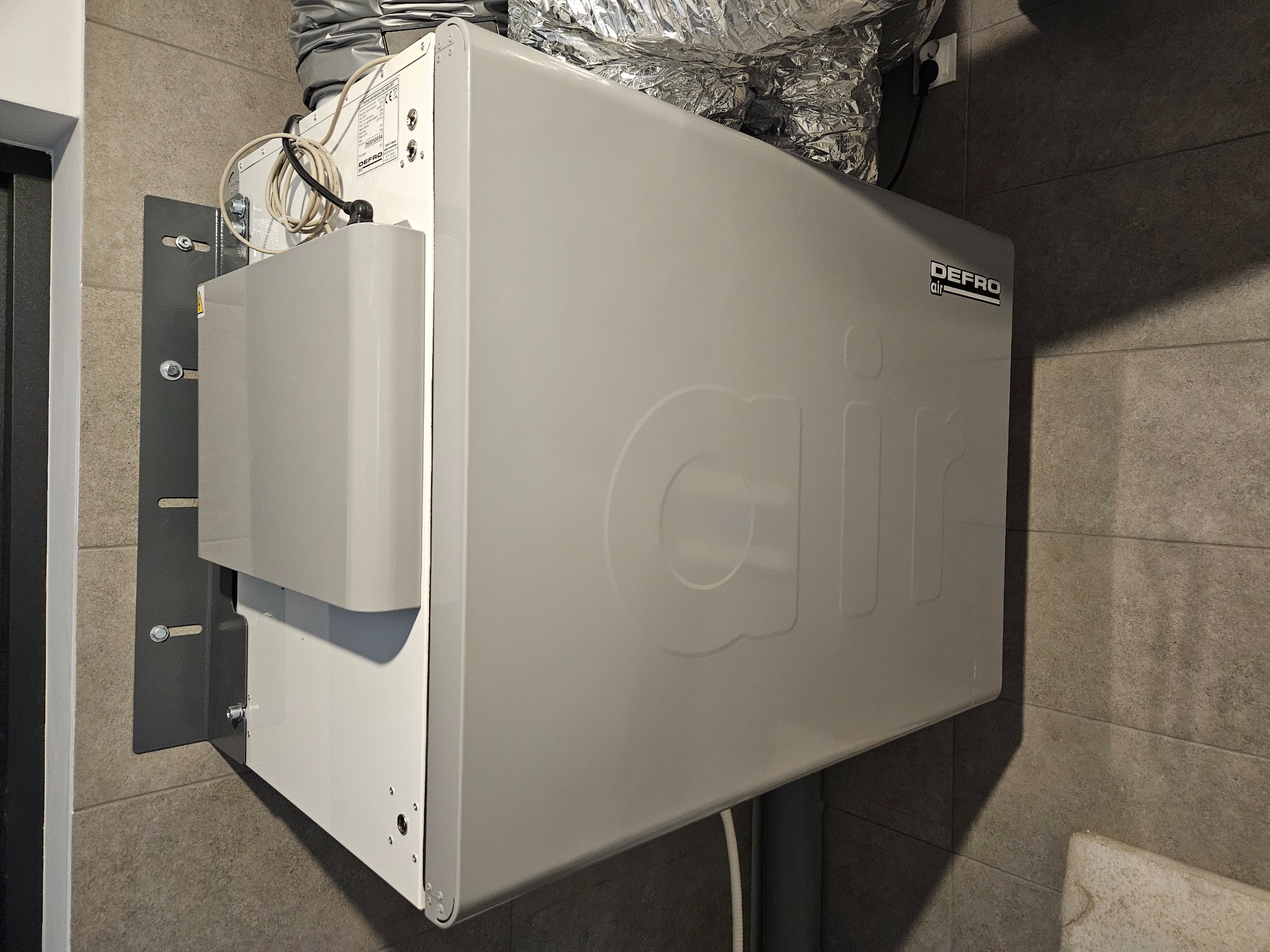 Zamontowany rekuperator Defro Air w pomieszczeniu z szarą płytką. Widoczne przewody wentylacyjne i uchwyty montażowe. Kątowe ujęcie, dobre oświetlenie.