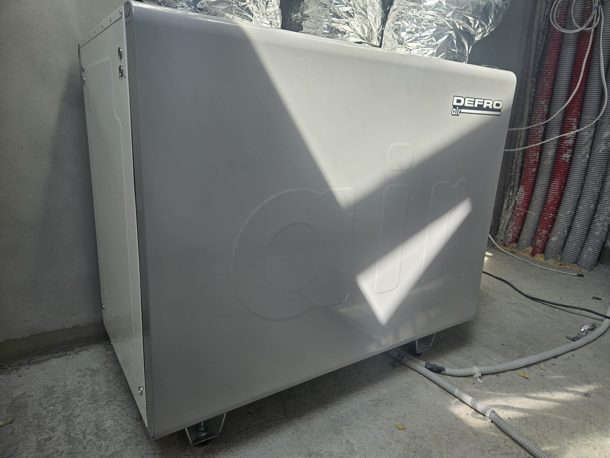 Biała jednostka rekuperacji DEFRO air na kółkach, widoczne rury wentylacyjne i przewody. Surowy beton na podłodze i ścianie. Oświetlenie dzienne.