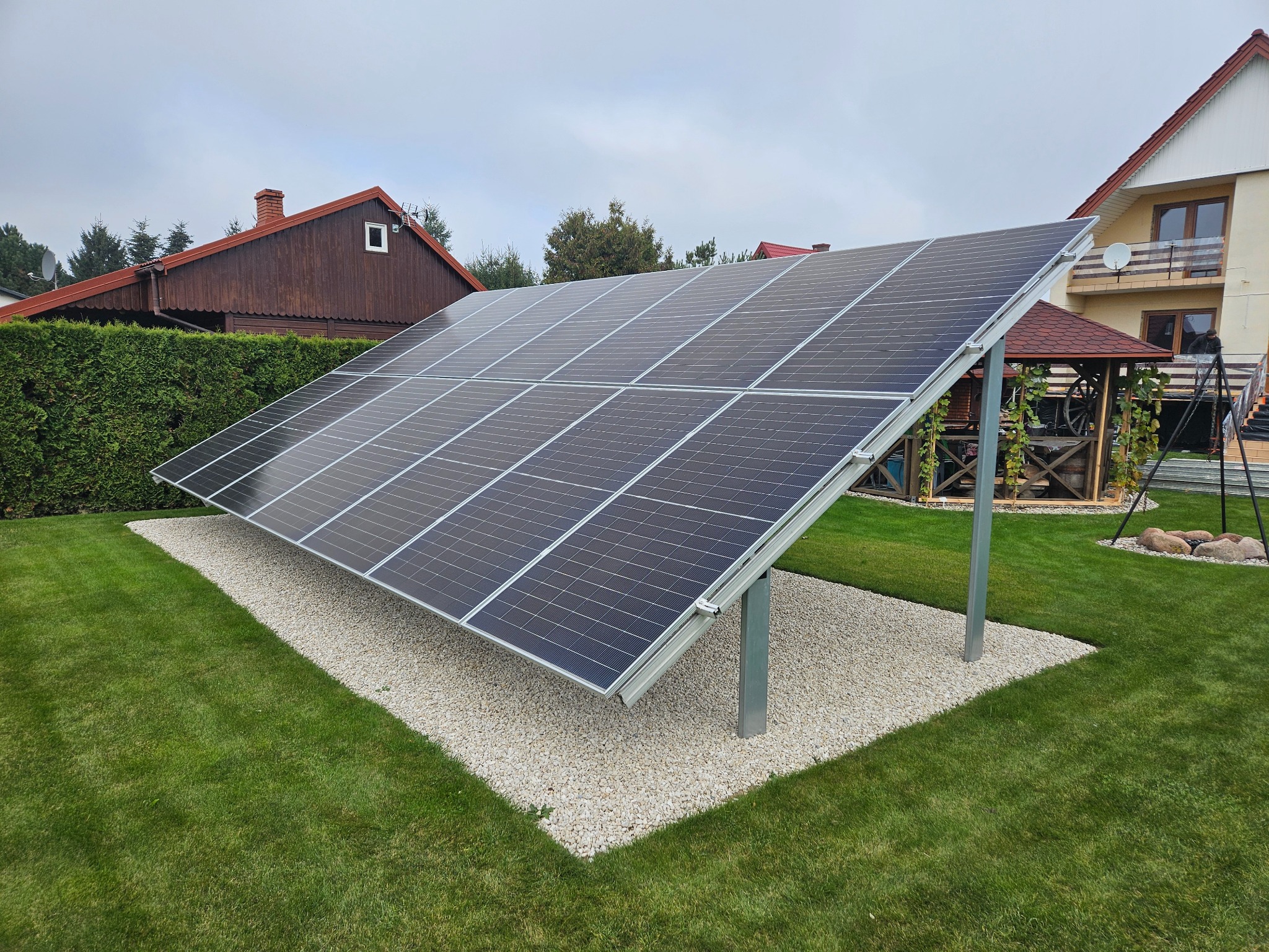 Panel fotowoltaiczny na trawniku przed domem jednorodzinnym. Instalacja oparta na metalowych słupkach, otoczona białym żwirem. Widok na budynek i altanę w tle.