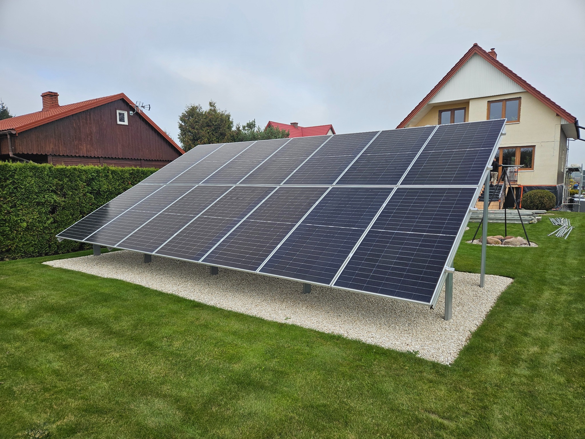 Panel fotowoltaiczny na trawniku przed domem jednorodzinnym. Instalacja na podłożu z jasnego żwiru. Widok na panele i domy w tle, pochmurne niebo.