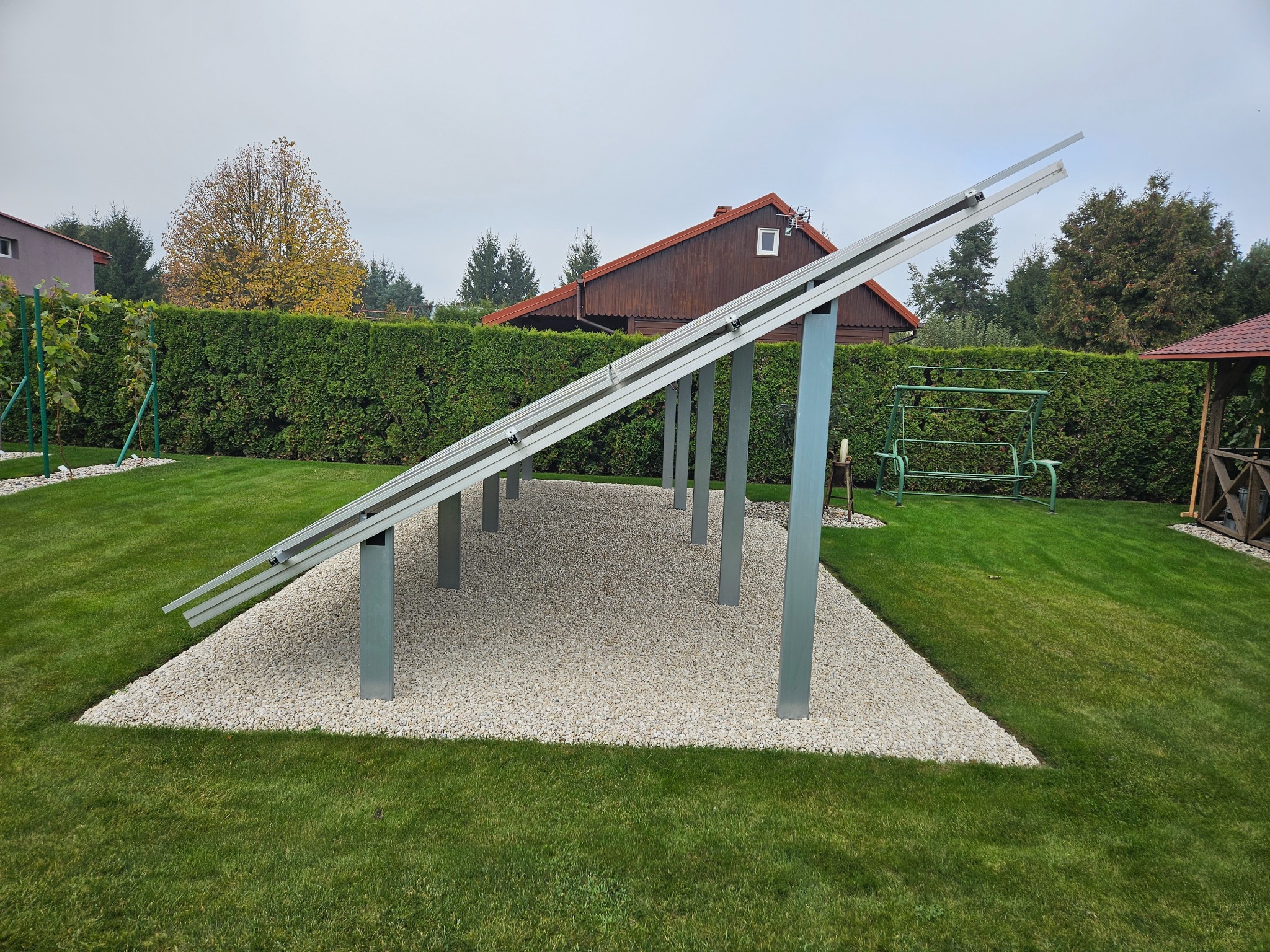 Stelaż pod panele fotowoltaiczne na posesji z trawnikiem i ozdobnym kamieniem. Widok z dołu, w tle dom z brązową elewacją i zielony żywopłot.