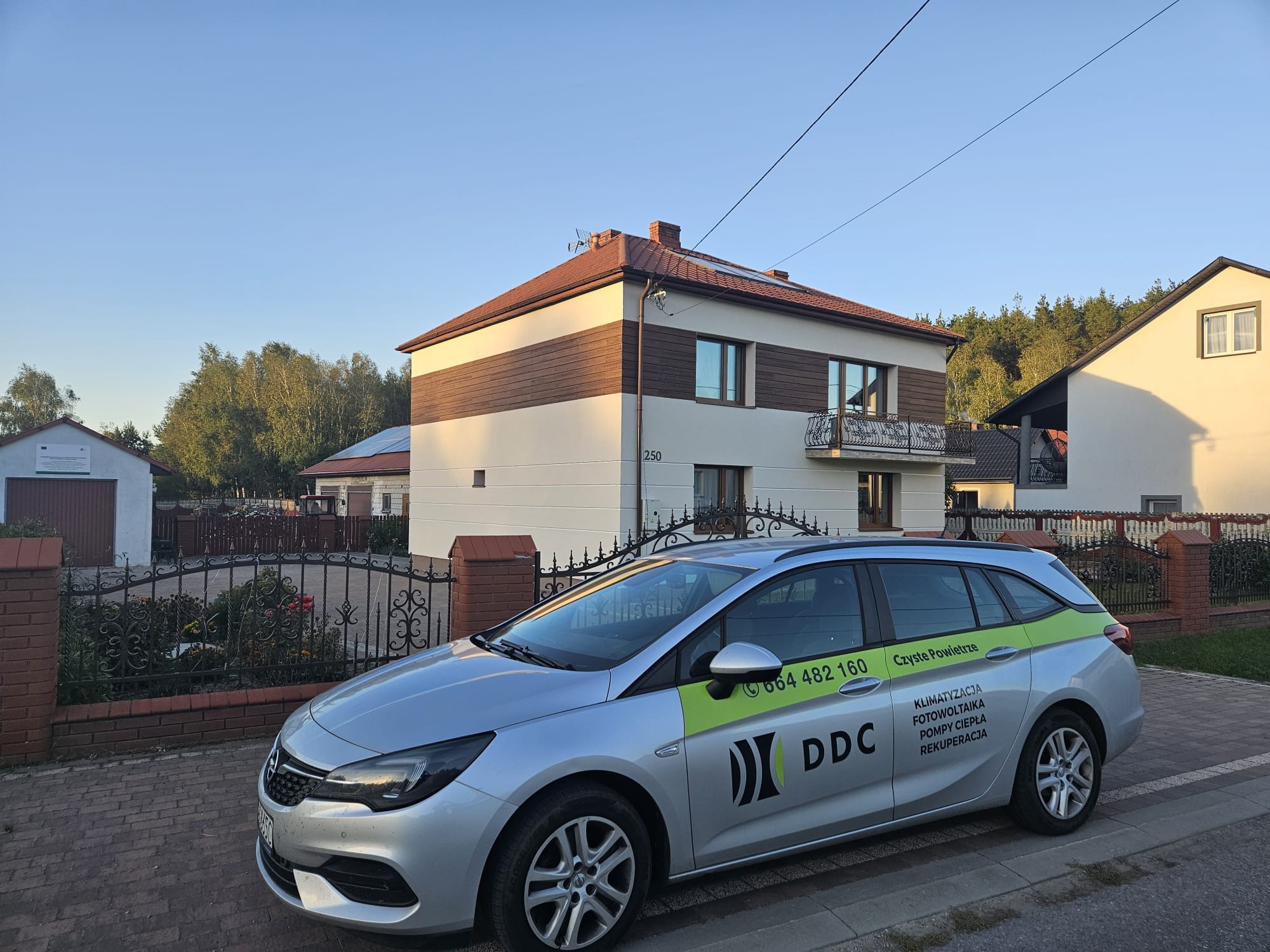 Dom z panelami słonecznymi na dachu, na pierwszym planie srebrny samochód z logo firmy oferującej klimatyzację, fotowoltaikę, pompy ciepła i rekuperację.