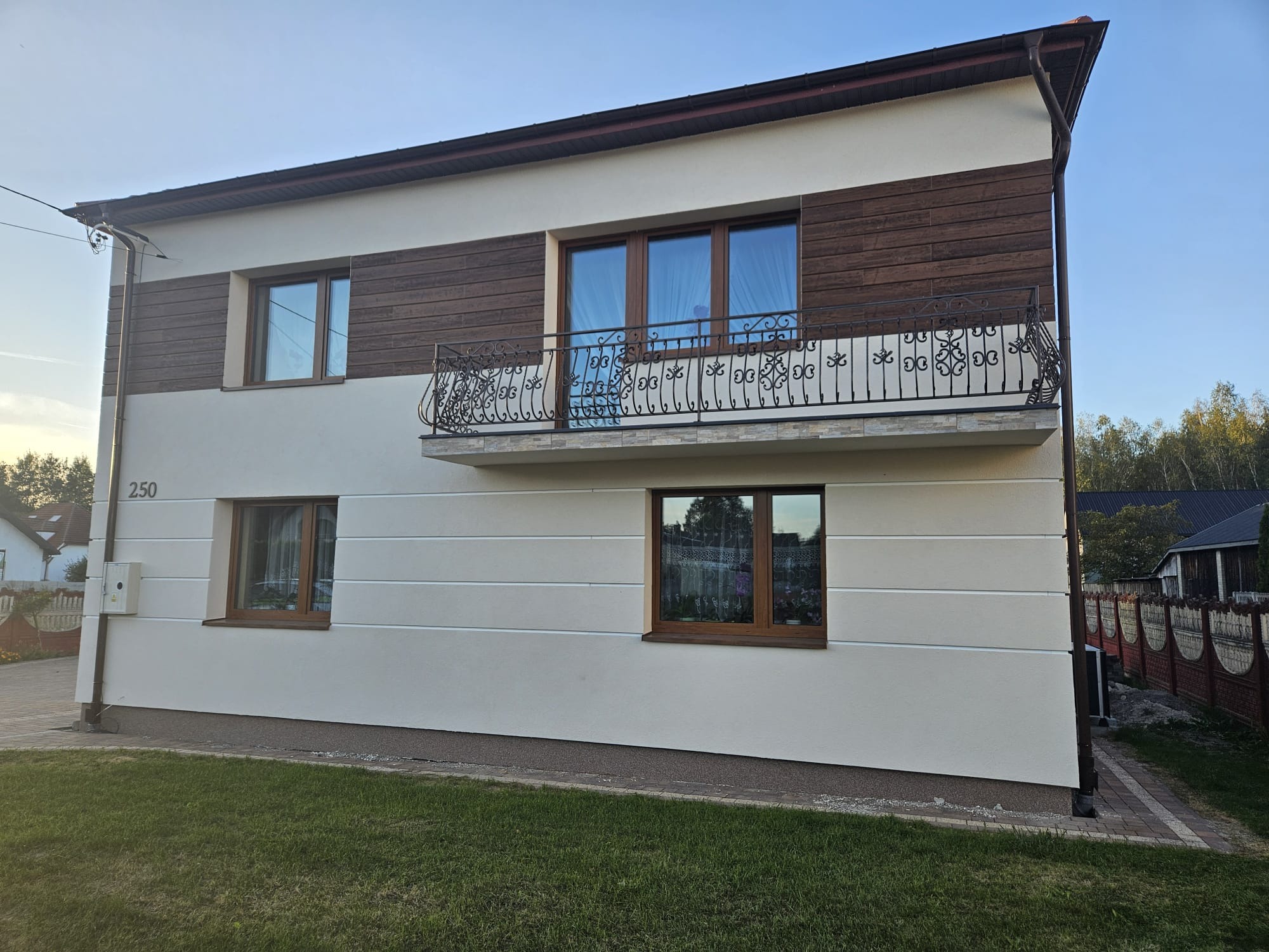 Dwupiętrowy dom z balkonem w Lublinie. Elewacja w odcieniach beżu i brązu, z drewnianymi panelami na piętrze. Ozdobna, metalowa balustrada balkonu. Widok z poziomu trawnika.