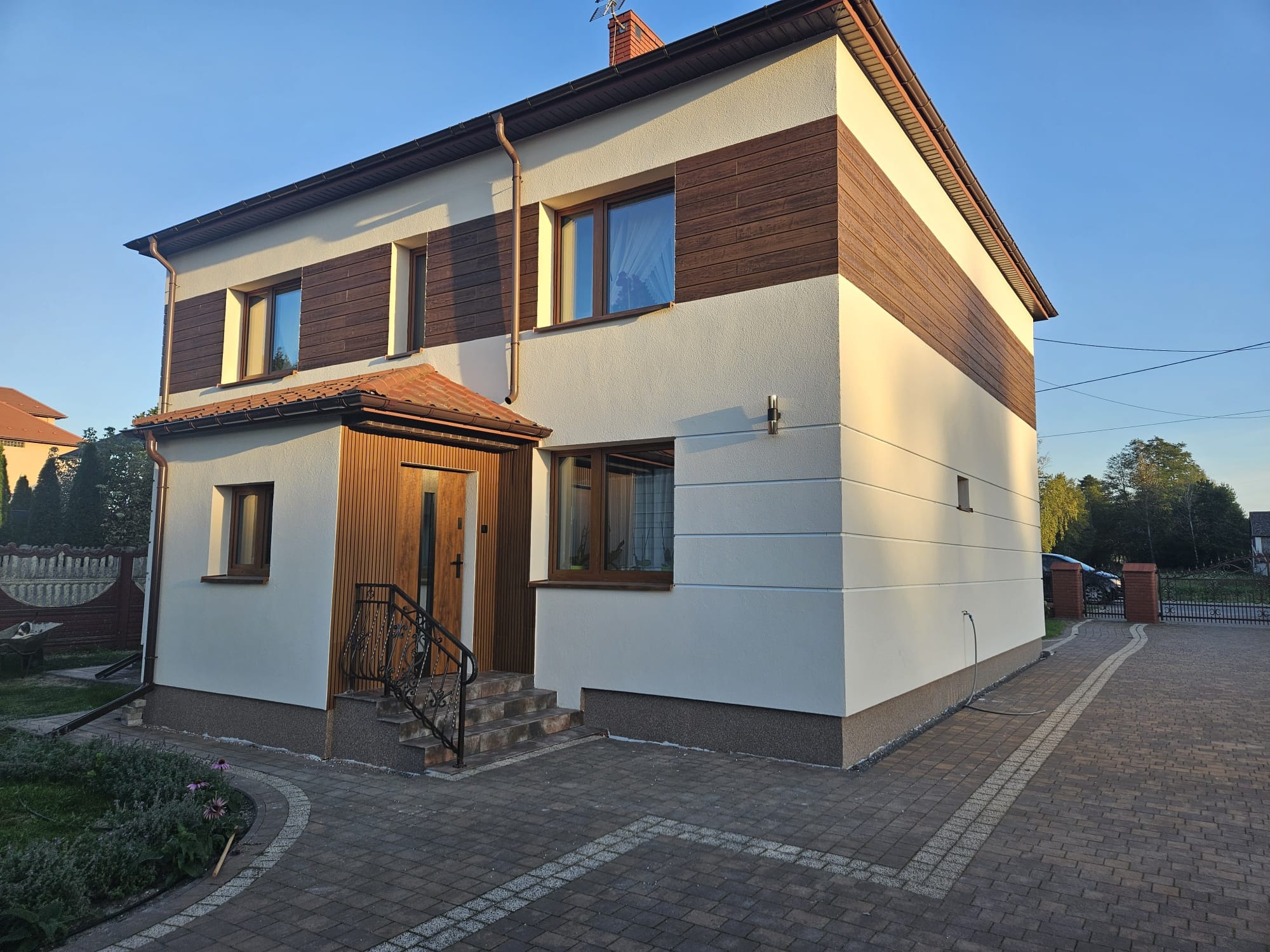 Dwukondygnacyjny dom z brązowymi panelami i jasną elewacją, widoczny z zewnątrz, z brukowanym podjazdem i metalową balustradą przy wejściu. Ujęcie w słoneczny dzień.