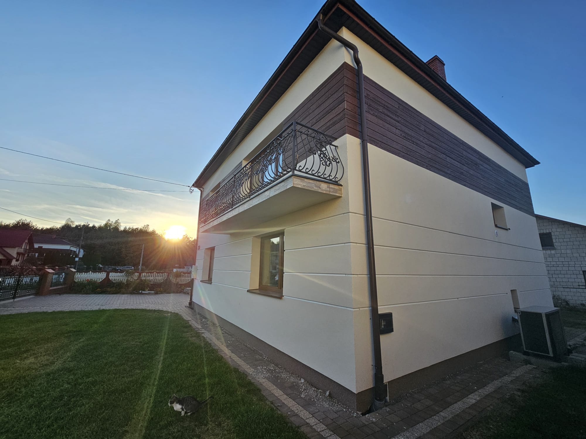 Elewacja domu jednorodzinnego z balkonem i brązowymi elementami dekoracyjnymi, widok z boku na tle zachodzącego słońca i zielonego trawnika z kotem.