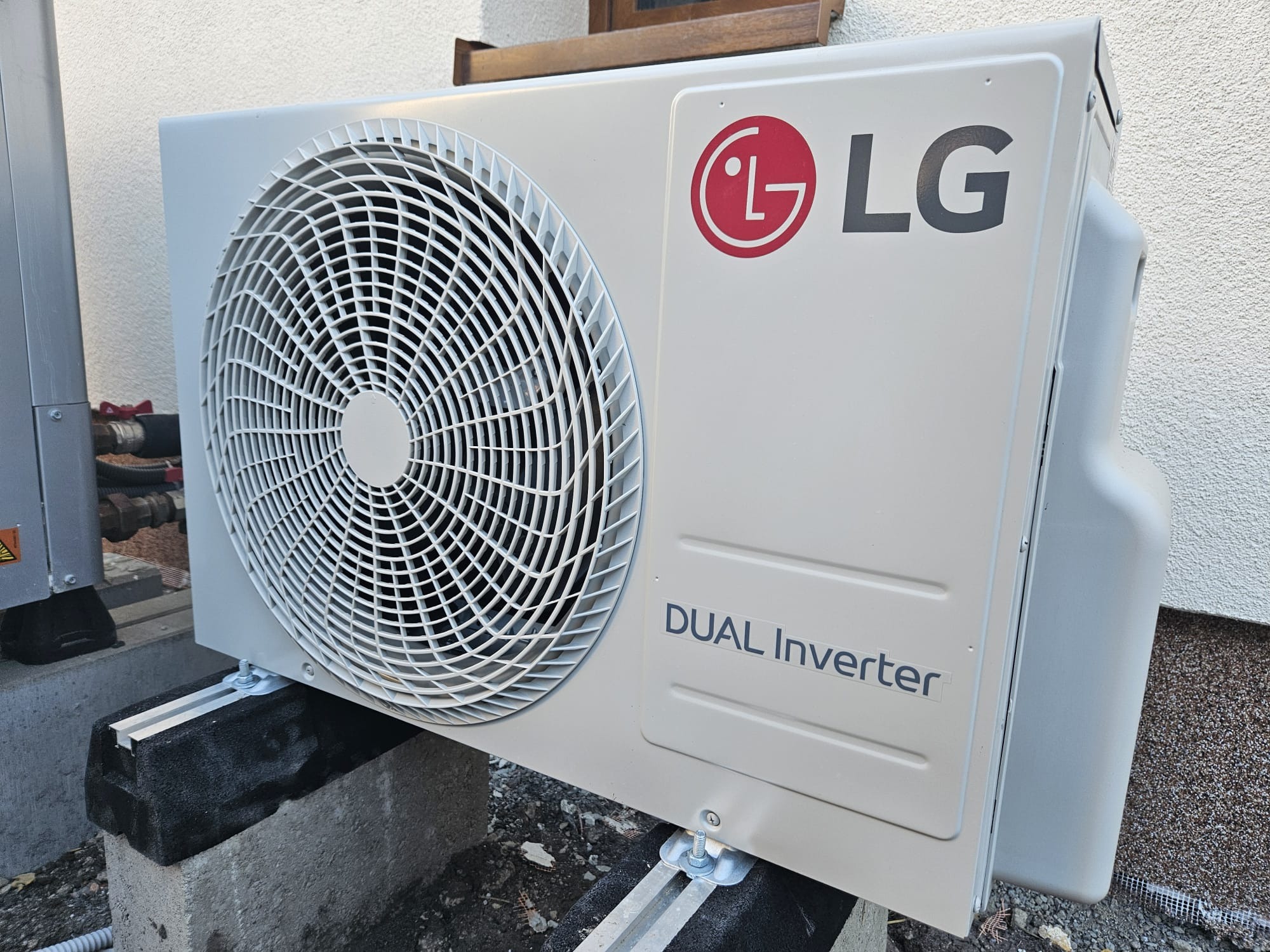 Zewnętrzna jednostka klimatyzacji LG Dual Inverter zamontowana na betonowych blokach przy ścianie budynku. Widoczne rury instalacyjne i wentylator jednostki.