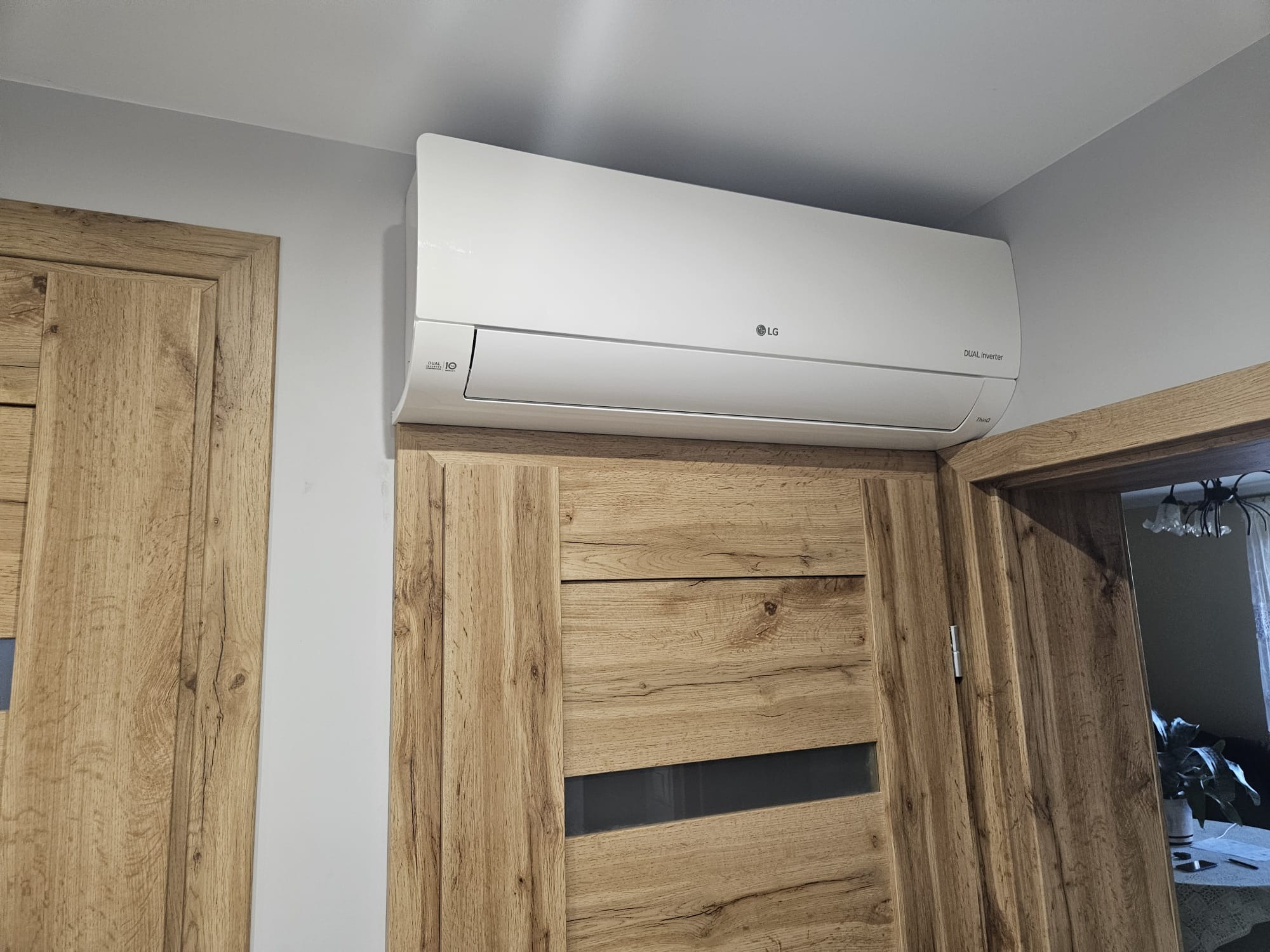 Biały klimatyzator LG Dual Inverter zamontowany nad drewnianą futryną drzwiową w jasnym wnętrzu. Widoczne detale urządzenia i fragmenty pomieszczenia.