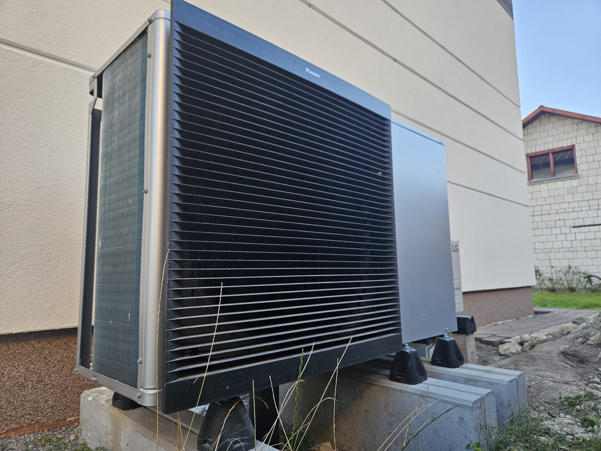 Zewnętrzna jednostka pompy ciepła Daikin, zamontowana na betonowych fundamentach przy ścianie budynku z widocznym fragmentem elewacji i sąsiedniego budynku.