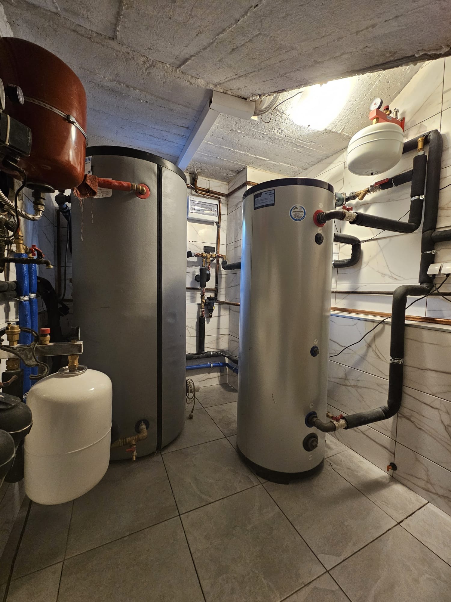 Dwa zbiorniki akumulacyjne ciepła w kotłowni, z widocznymi rurami i armaturą. Jeden zbiornik w szarej obudowie, drugi z brązowym elementem na górze.