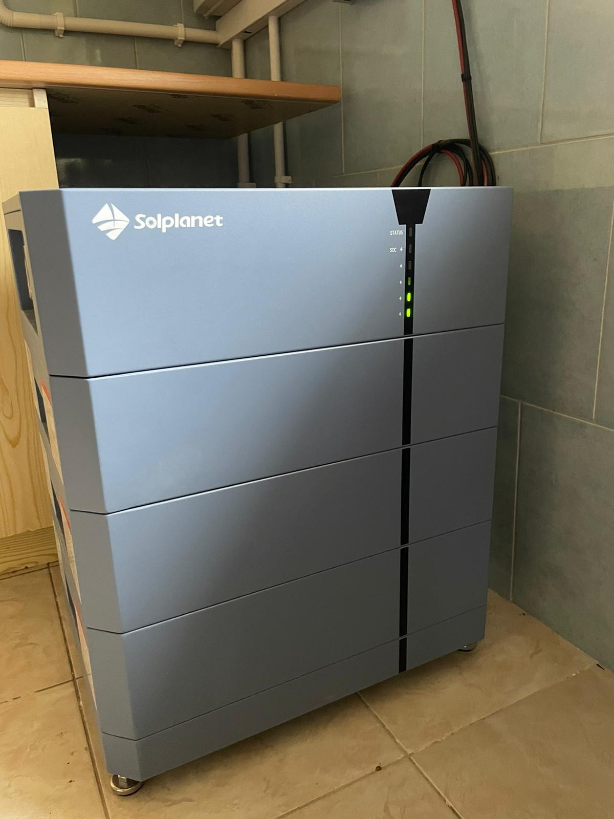 Szary akumulator energii Solplanet z diodami LED stanu na tle beżowych płytek i drewnianej półki. Widoczne przewody. Minimalistyczny design.