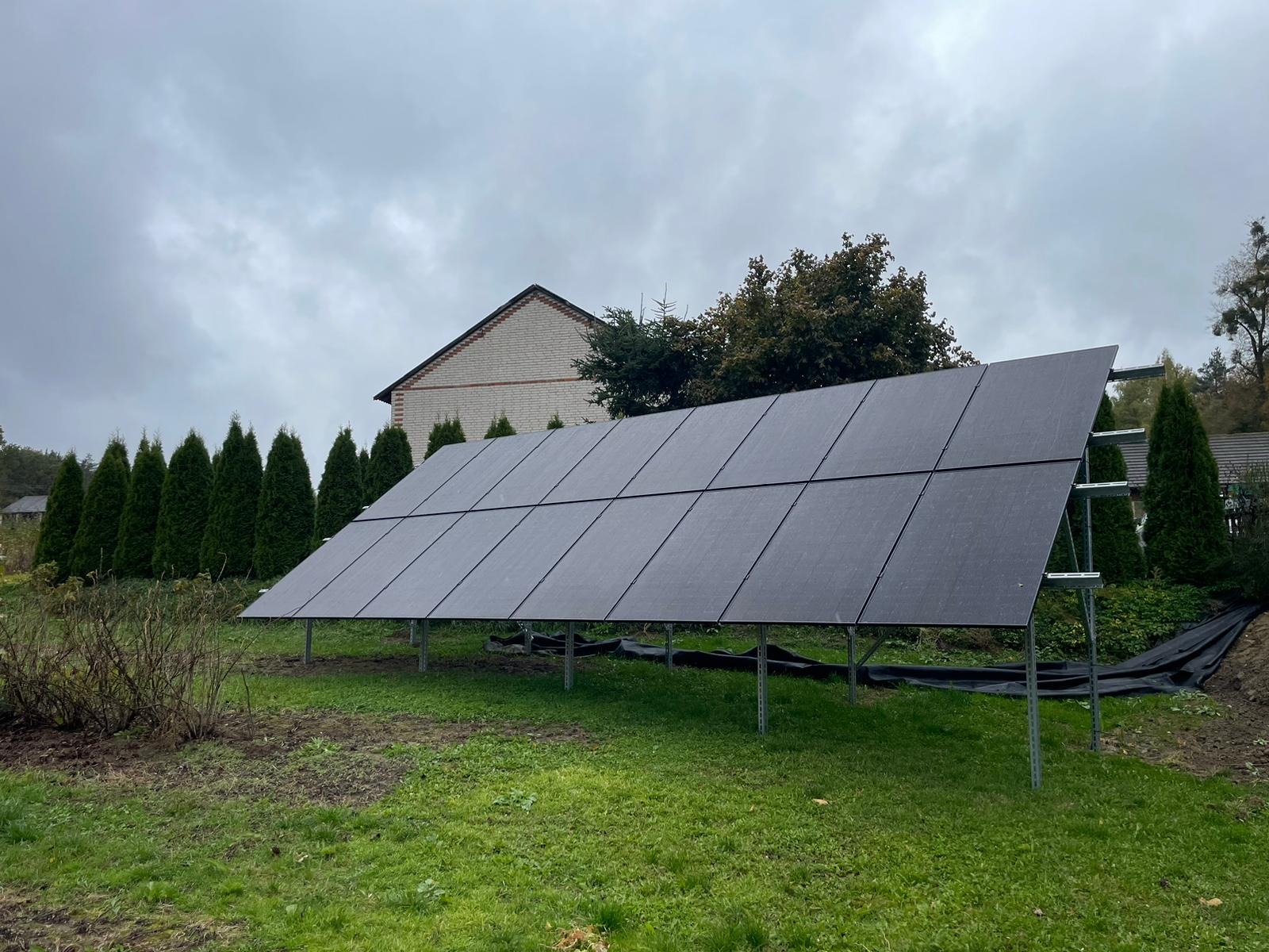 Panel fotowoltaiczny na trawie, widok z boku, pochmurne niebo w tle. Instalacja paneli na ziemi, widoczne podpory i elementy montażowe. Dom w oddali.